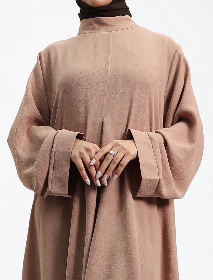 A woman wearing a loose, high-neck, long-sleeved abaya in a dusty rose-brown color, paired with a black hijab. The photo is cropped from the neck down, showing her hands with manicured nails and a ring, set against a plain white studio background.