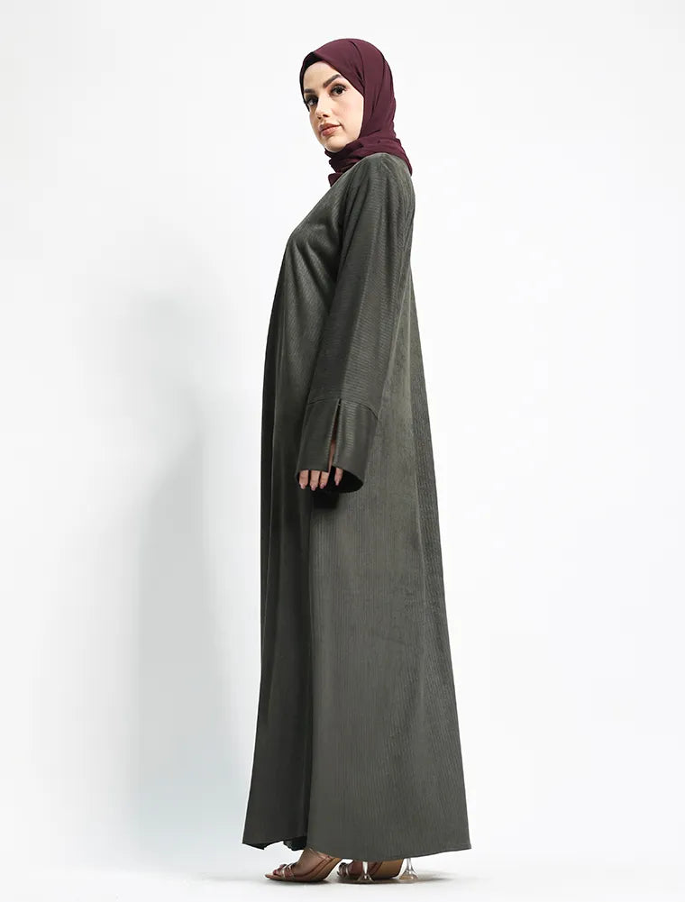 A woman wearing a dark gray abaya and a deep maroon hijab, standing in profile against a plain white studio background.