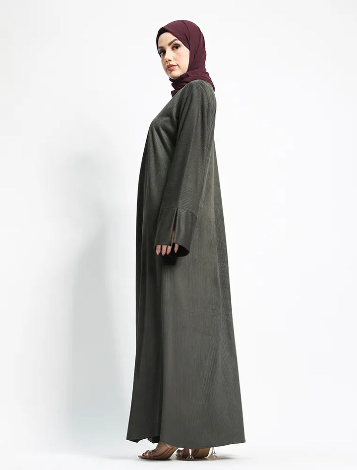 A woman wearing a dark gray abaya and a deep maroon hijab, standing in profile against a plain white studio background.