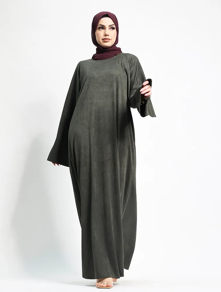A woman modeling a long, loose-fitting dark olive green abaya dress with wide sleeves, paired with a deep plum hijab, standing against a plain white studio background.