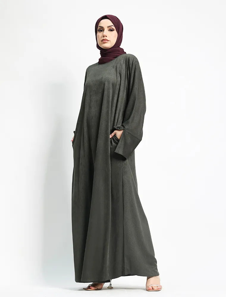 A woman wearing a dark gray long-sleeved abaya with a matching headscarf, standing against a plain white studio background.