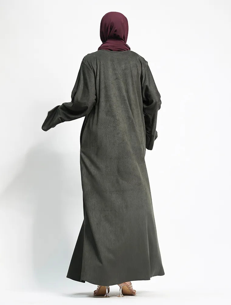 A woman viewed from behind wearing a long, loose-fitting dark gray abaya and a deep plum hijab, standing against a plain white studio background.
