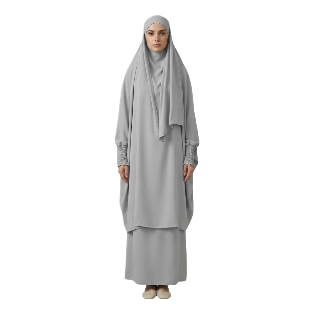 A woman modeling a light gray modest outfit consisting of a long abaya with lace-trimmed sleeves, a matching skirt, and a hijab, set against a plain white background.