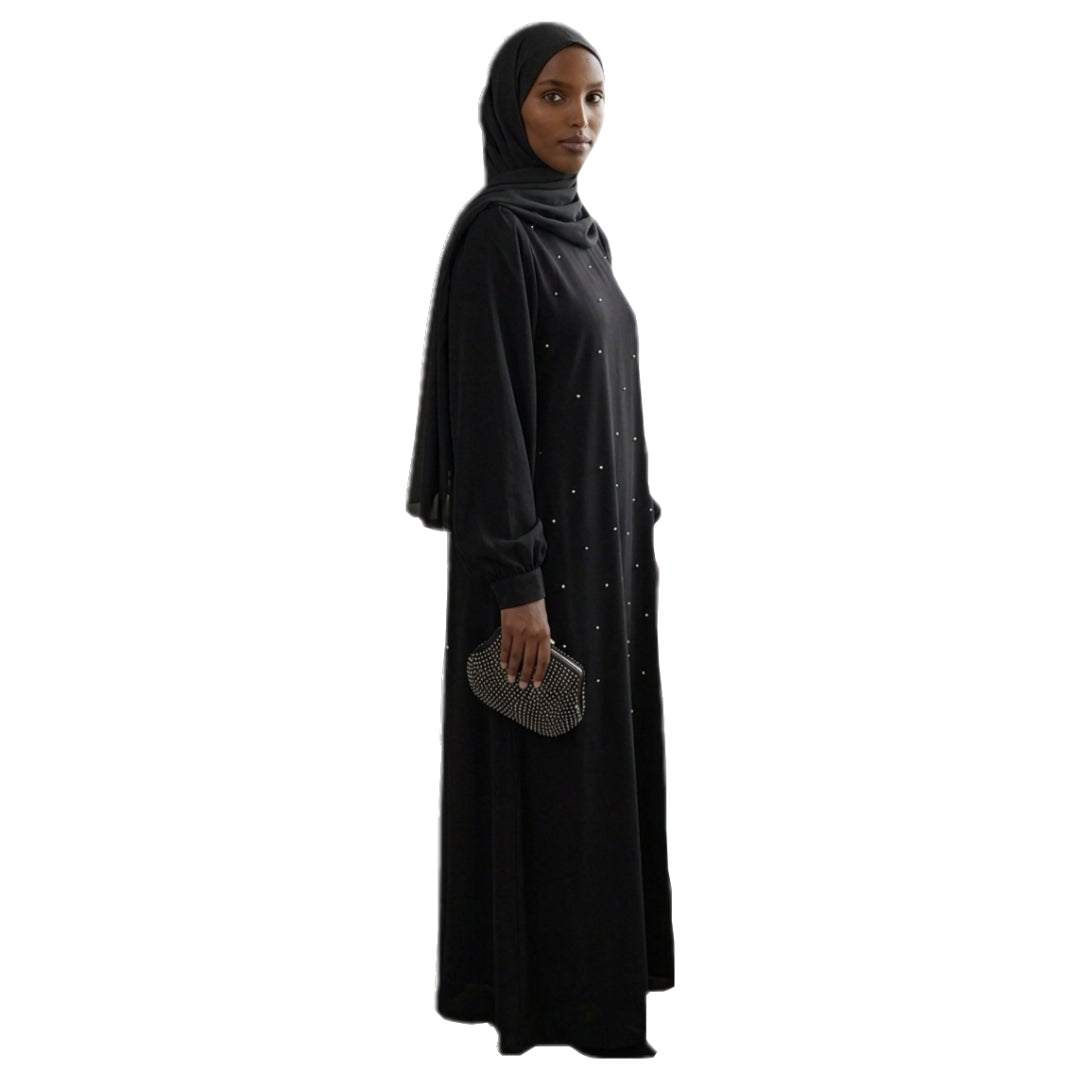 A woman wearing a black modest long-sleeve abaya dress with small silver rhinestone embellishments, a matching black hijab, and holding a small silver clutch, shown against a plain white background.