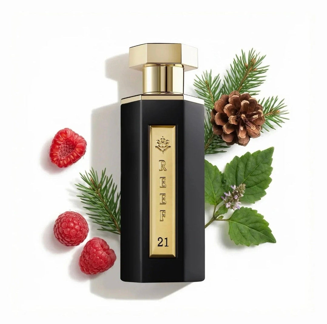 A black rectangular perfume bottle with a gold cap and gold vertical lettering reading 'reef 21' is centered on a white background, surrounded by holiday greenery and red raspberries. The photo appears to be a product listing image, though it is shown within a phone app interface with black bars around it.