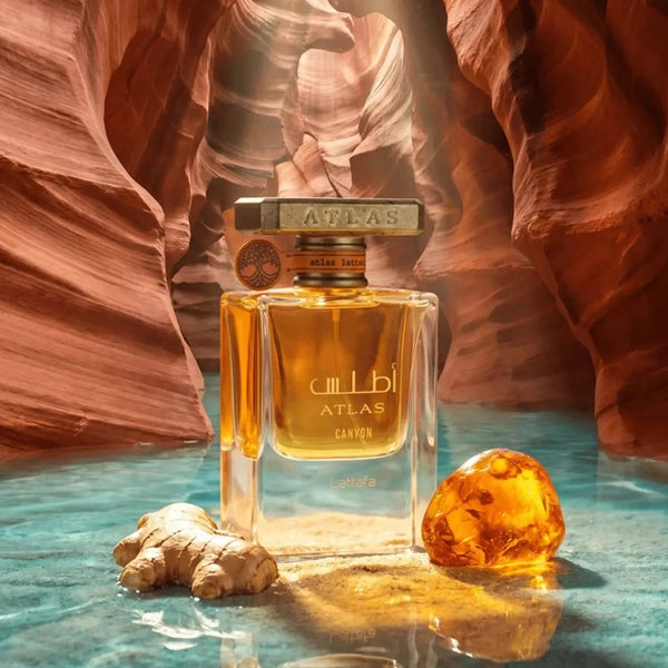 Atlas Canyon perfume bottle displayed in a canyon-like setting with turquoise water, a ginger root, and an amber stone. The bottle is clear glass with golden-amber liquid and a metallic cap engraved with 'ATLAS'.